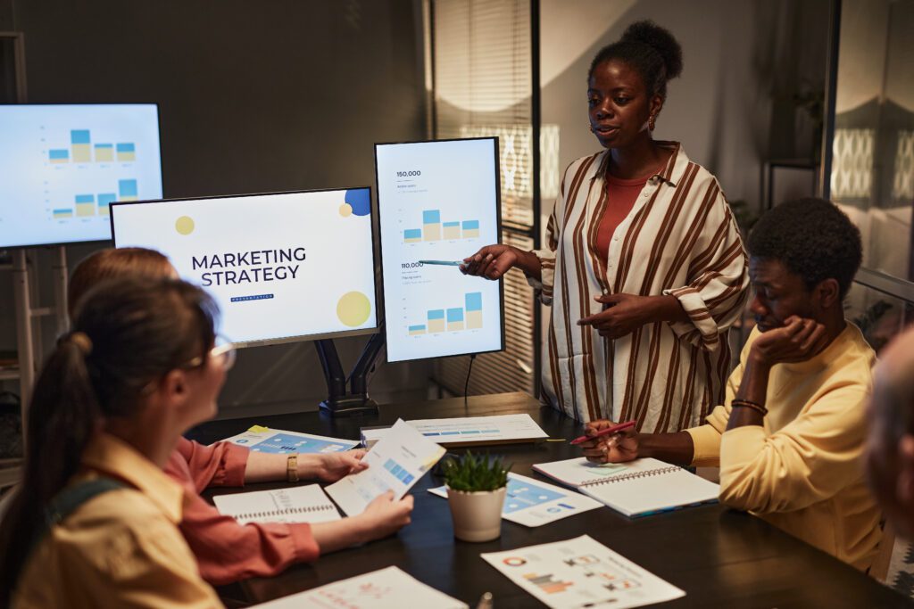 Web Design - Portrait of young black woman giving presentation on marketing to team in dimly lit office